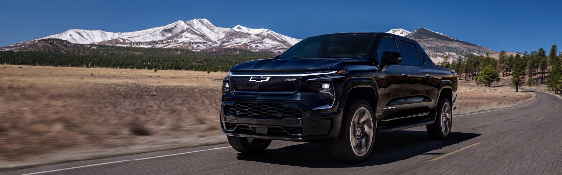 A black Chevrolet Silverado truck driving on a scenic road with snow-capped mountains in the background.