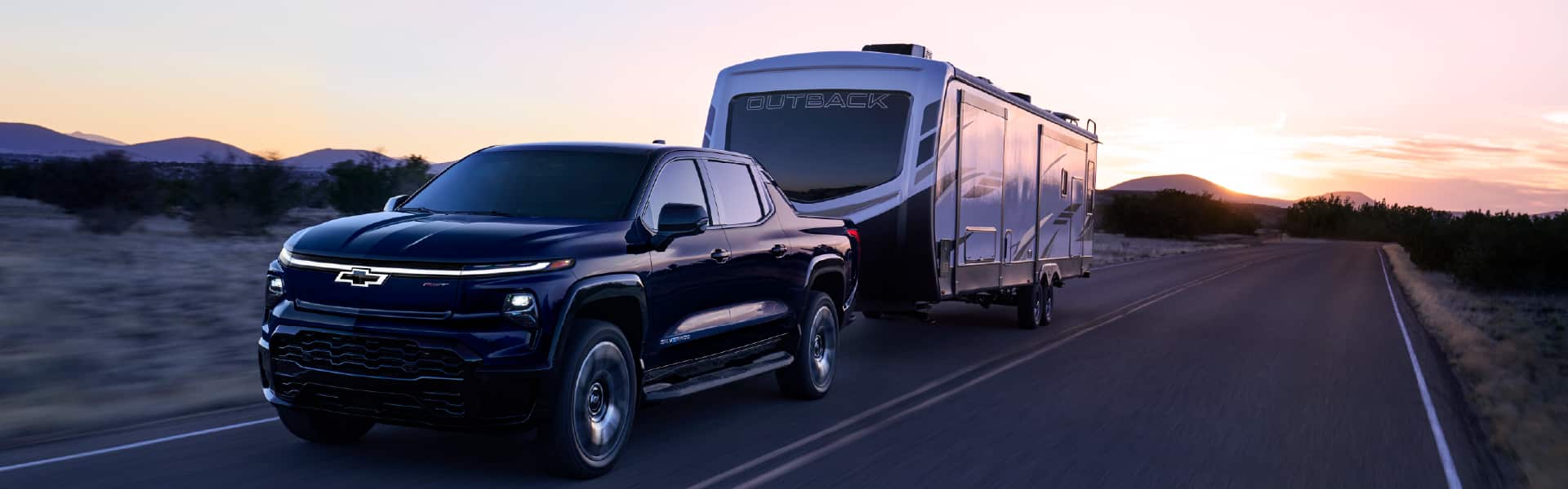 A blue Chevrolet truck towing a camper trailer on an open road at sunset.