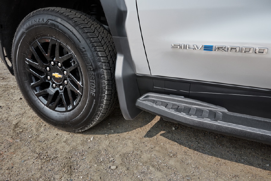 Close-up of a Chevrolet Silverado tire and step with logo, parked on gravel.